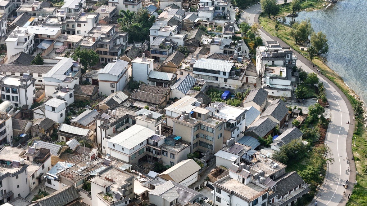 Aerial view of a dense lakeside village of tiled roofs beside a curving road along the Erhai Lake shore near Xizhou.