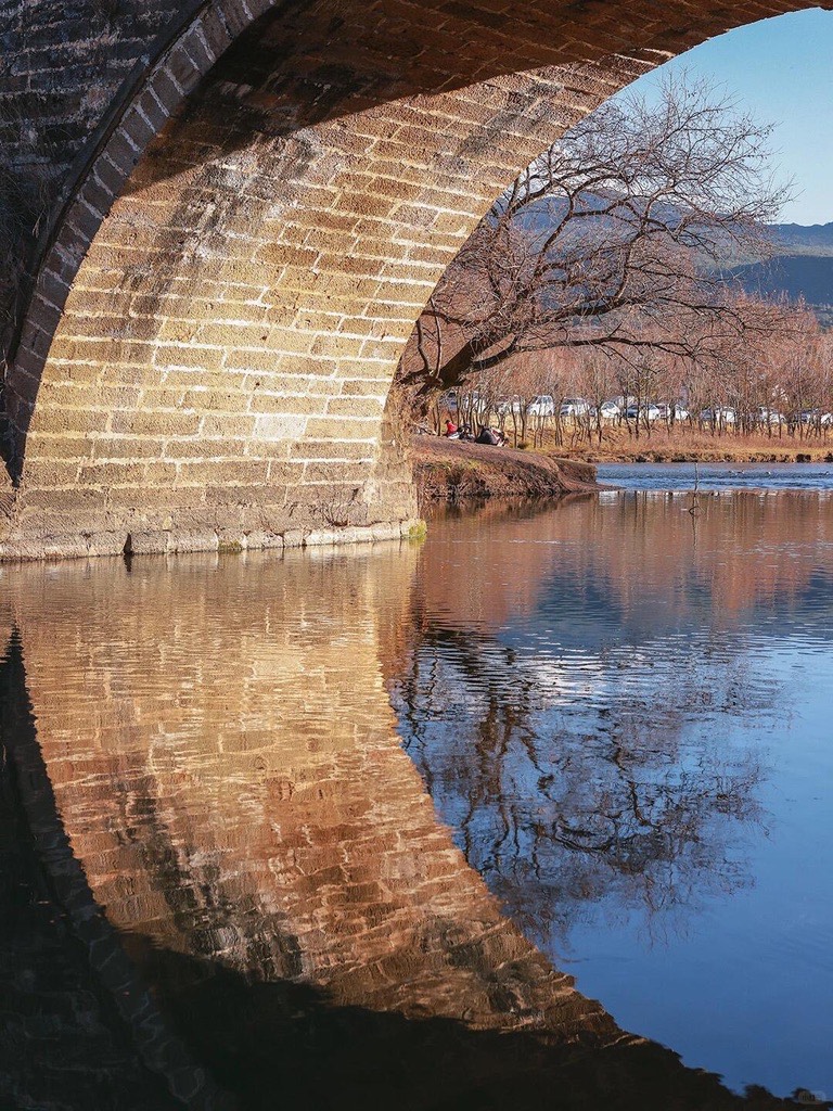 Old stone arch bridge and bare riverside tree mirrored in the calm water at Shaxi Ancient Town.