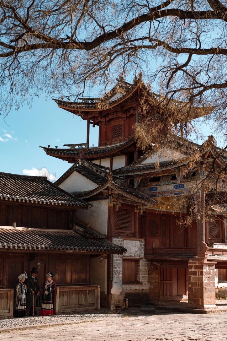 Weathered tiered timber theatre pavilion in Shaxi ancient town, with locals in traditional dress on the caravan-trail square near Dali.