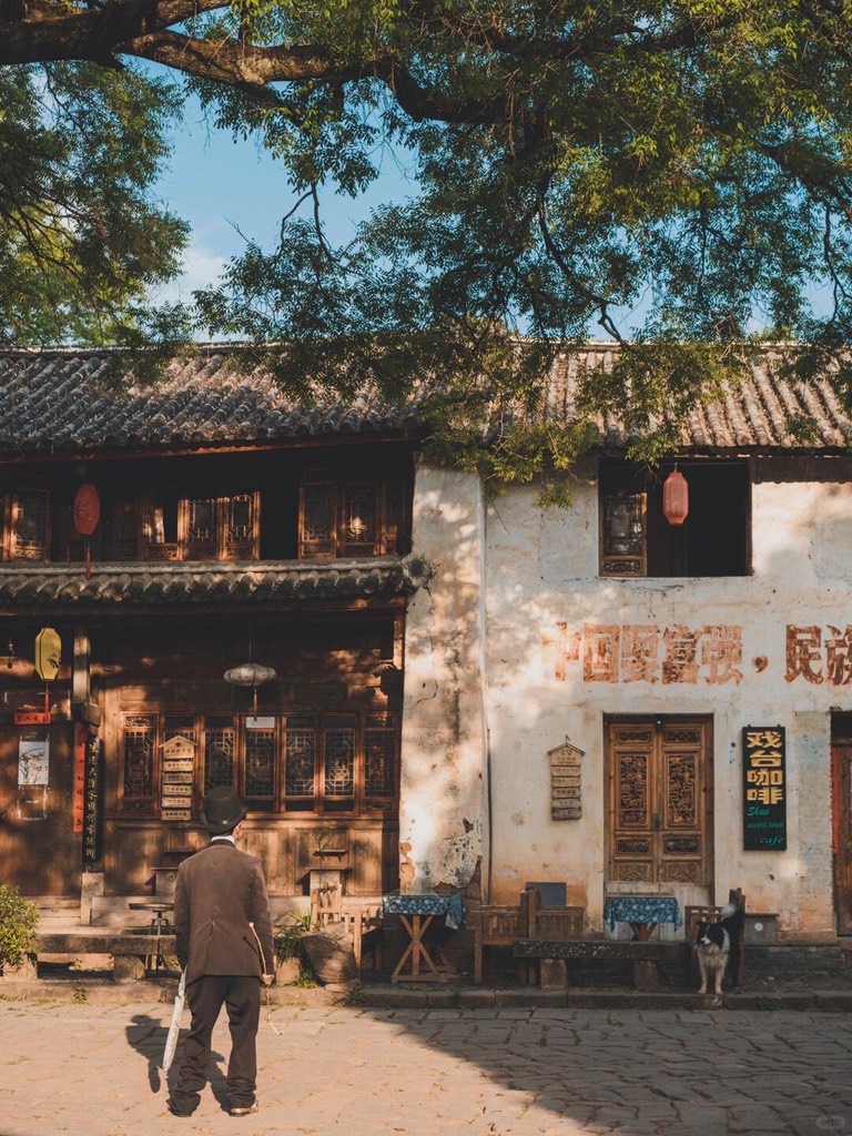 Elderly man crossing a sunlit cobbled square before weathered timber shophouses in Shaxi Ancient Town.