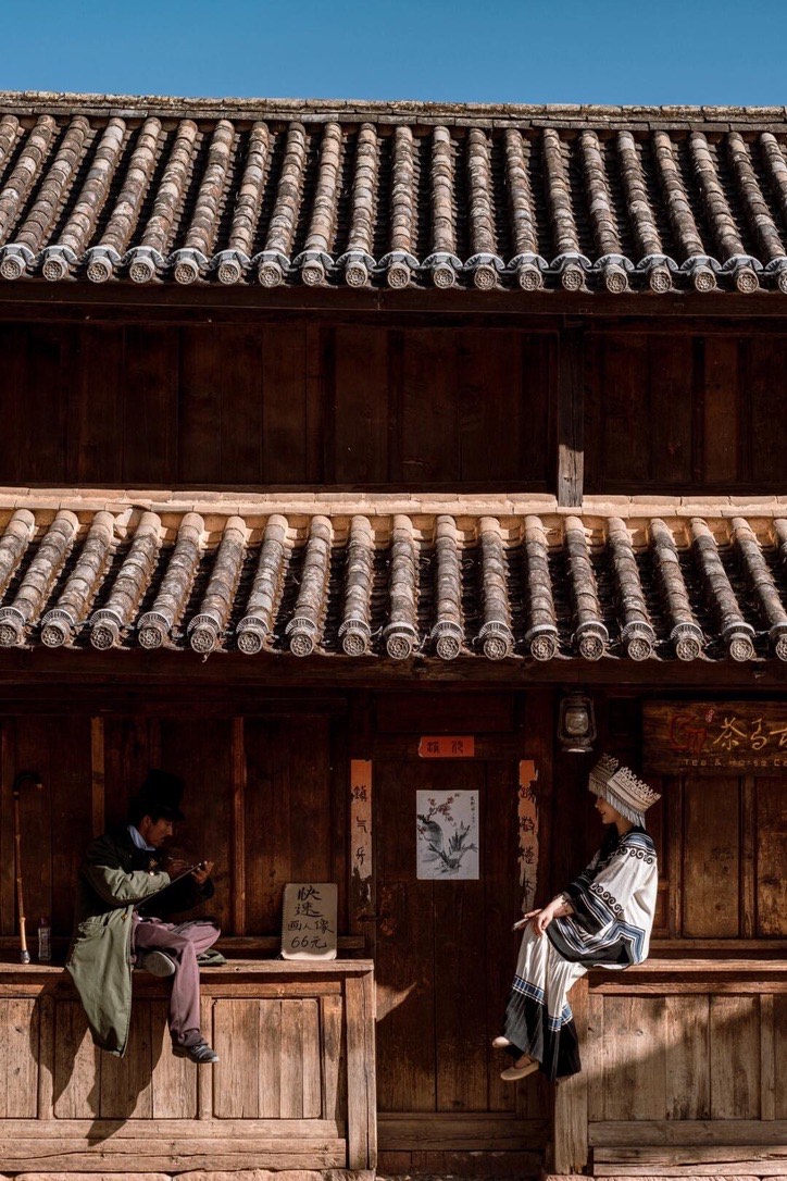 A woman in embroidered ethnic dress sits on a wooden storefront under tiled eaves as a man sketches in Weishan Ancient Town.