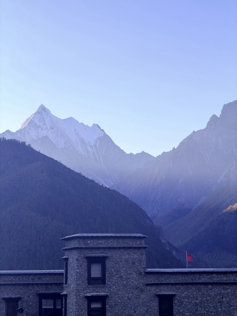 Snow-capped peaks glowing at sunrise above a traditional Tibetan stone lodge in Yading village.