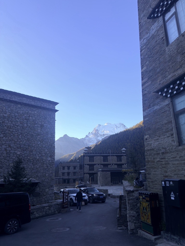 Stone Tibetan guesthouses and parked cars in Yading village with a snow-capped peak in the morning light.