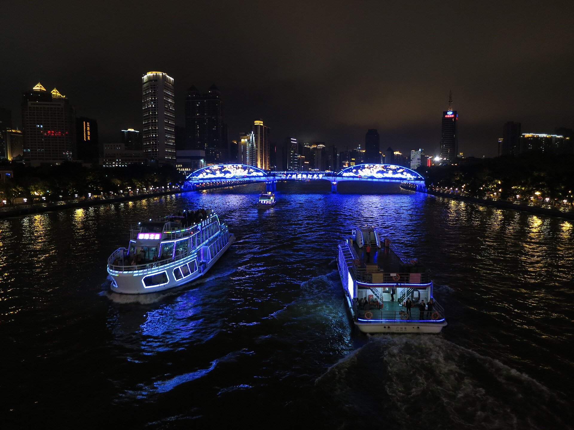Pearl River night-cruise boats (珠江夜游) gliding past a blue-lit bridge with the Guangzhou skyline behind