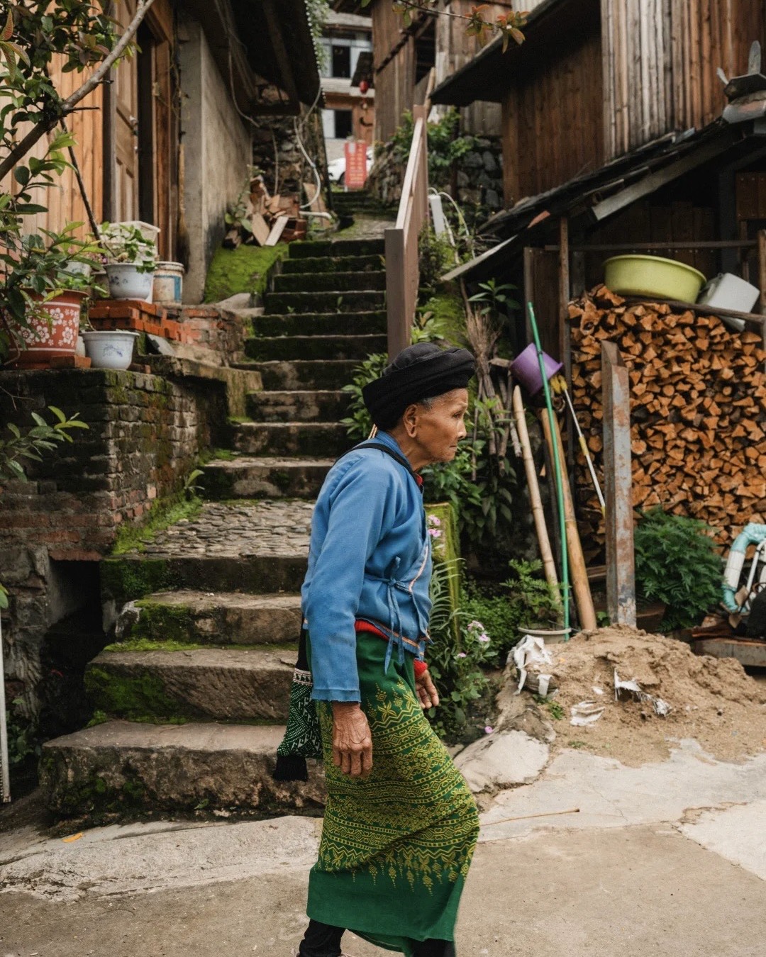 Elder woman in a blue jacket and green embroidered skirt walks a village lane past mossy stone steps and stacked firewood on Jingmai Mountain