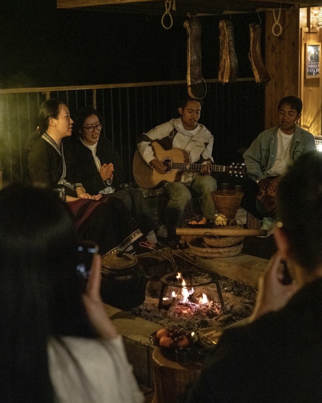 Guests gather around an indoor firepit with guitar and cured meats hanging above at a Jingmai Mountain lodge.