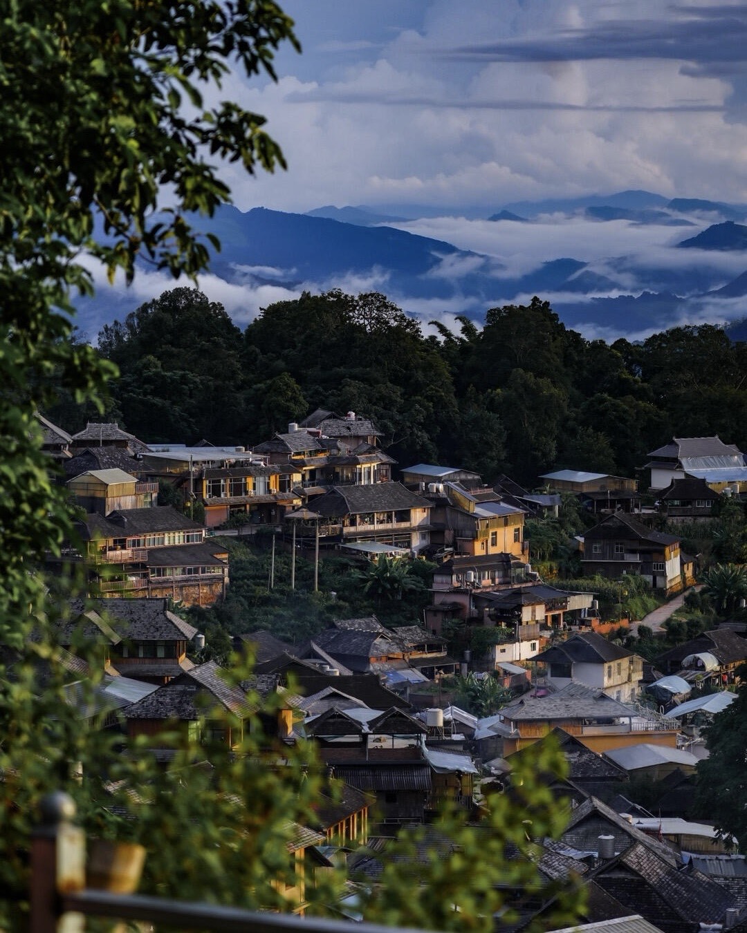 Cluster of tiered village houses on a wooded hillside with misty mountain ridges beyond at dusk on Jingmai Mountain.