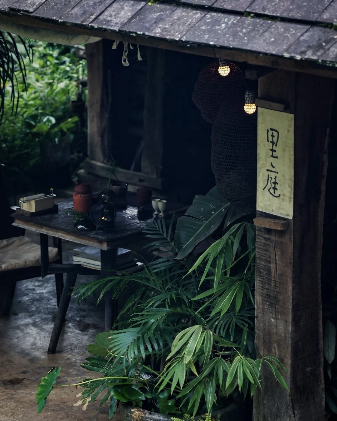 Rustic timber tea house with a lit lantern, teaware on a wooden table and lush green plants on Jingmai Mountain.