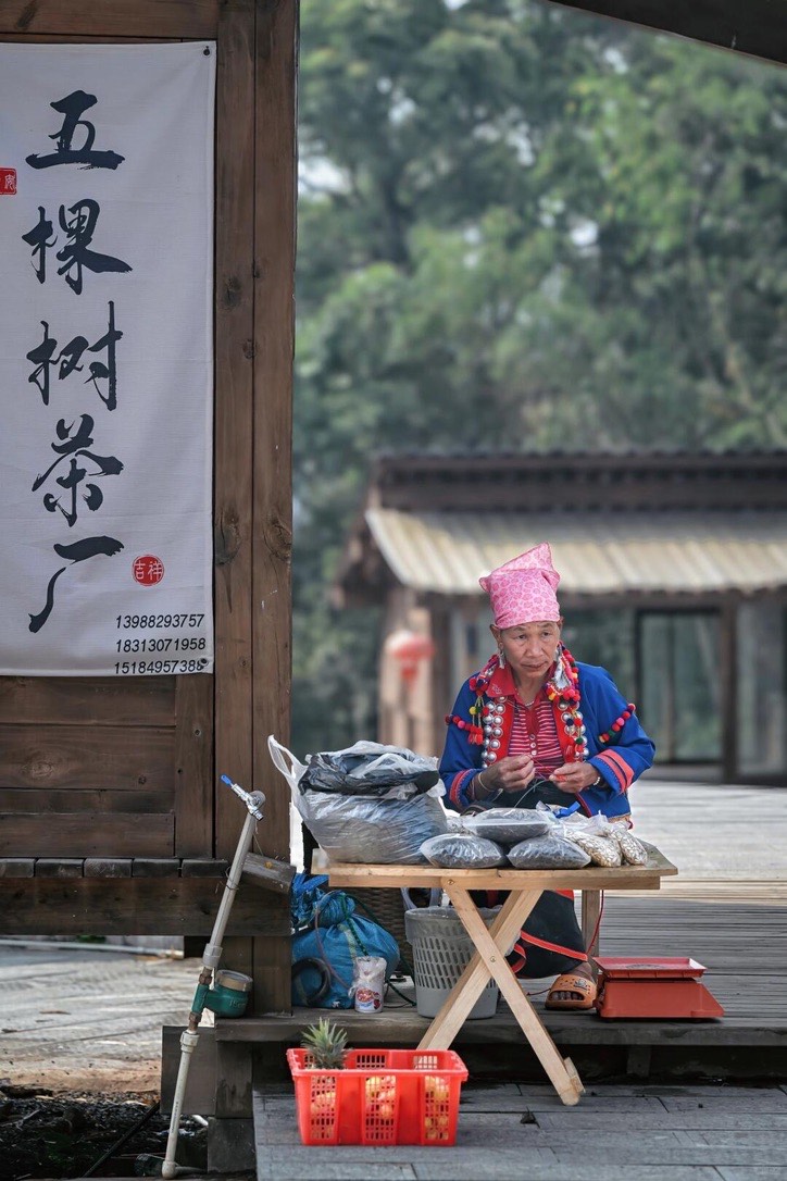 Woman in traditional dress and pink headdress sorting tea at a small wooden stall by a village boardwalk on Jingmai Mountain.