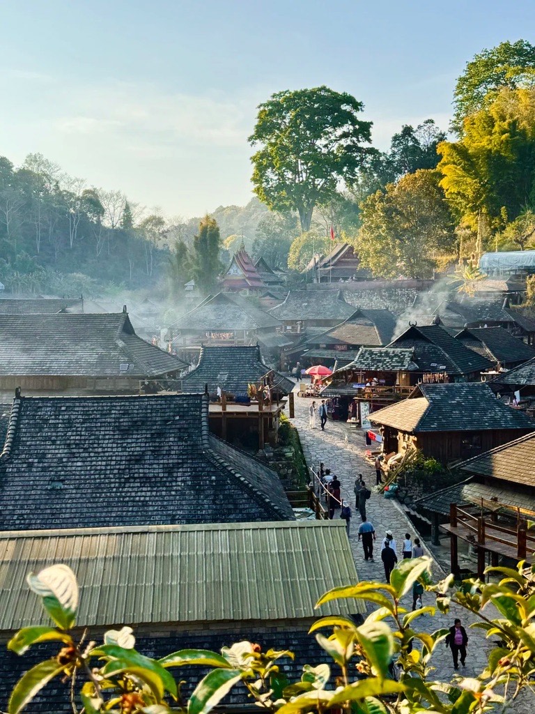 Elevated view over wood-shingled village rooftops and tea plants with morning mist and visitors on a stone path on Jingmai Mountain