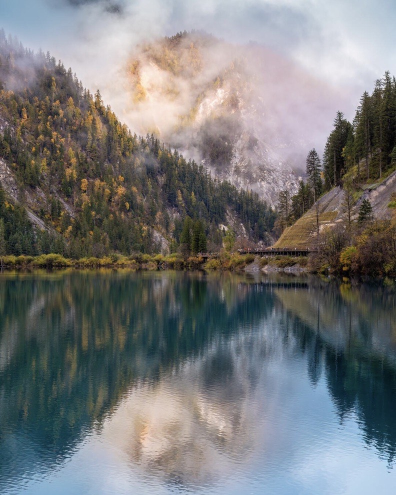 Misty morning light over a turquoise Jiuzhai Valley lake, pine forest and rock faces reflected in still water
