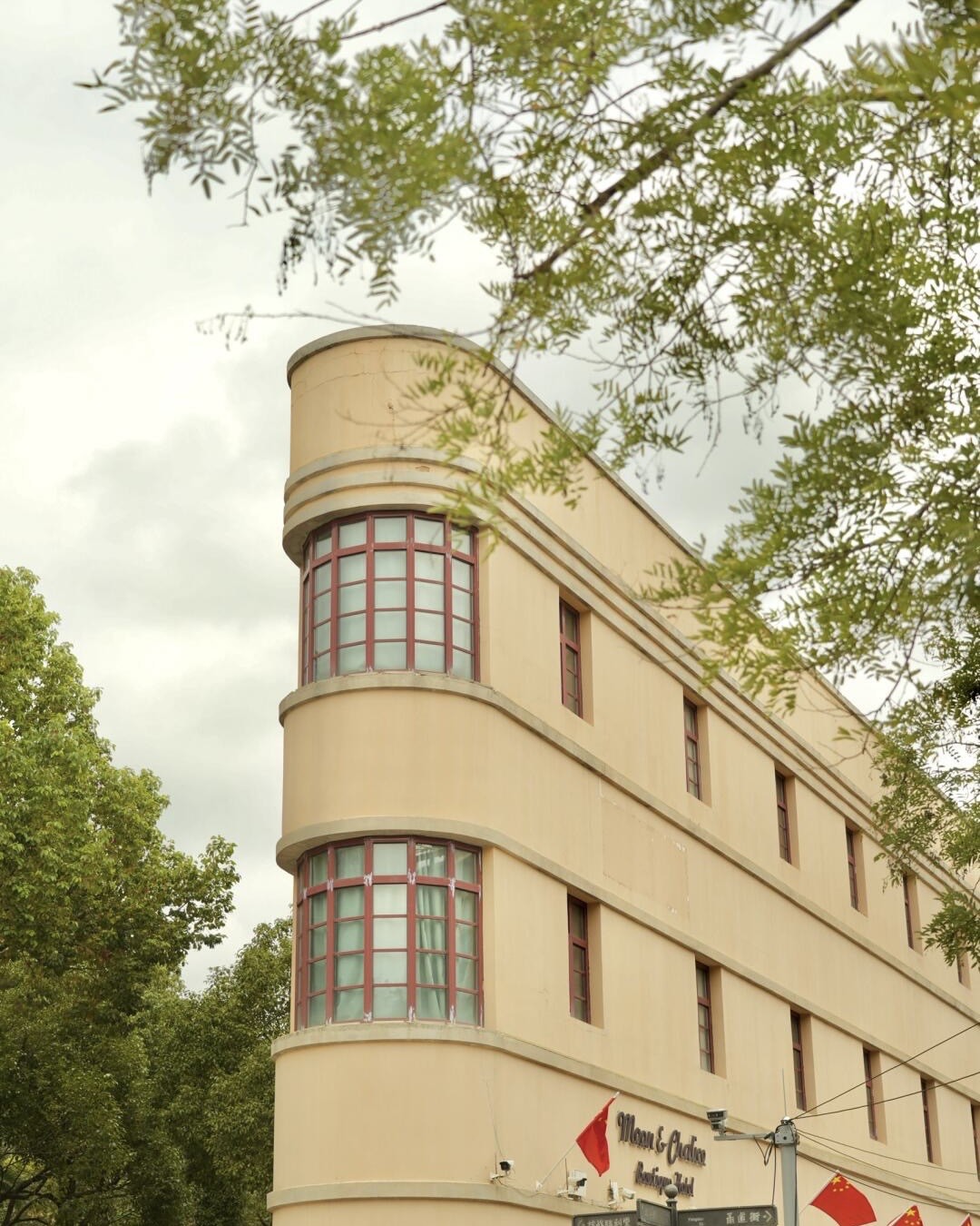 A curved cream-coloured Art Deco corner building with red-framed windows stands among leafy trees in Kunming.