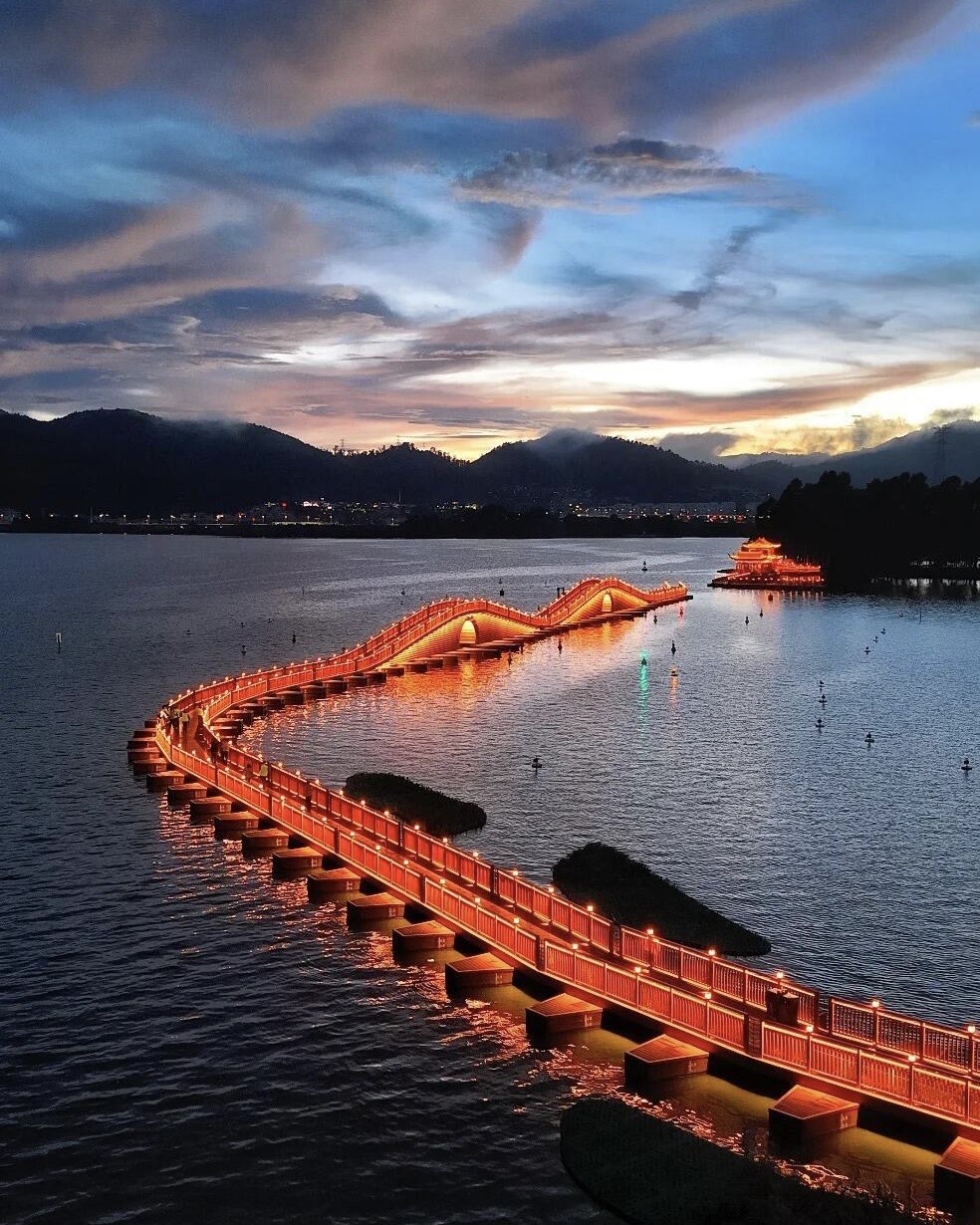 A winding illuminated boardwalk bridge glows orange across Dianchi Lake at dusk below West Mountain in Kunming.