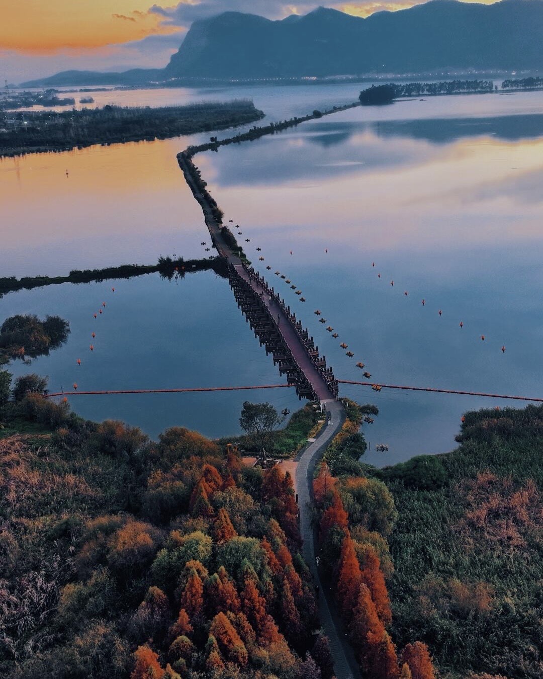 Aerial view of a wooden boardwalk crossing Dianchi Lake at sunset with West Mountain silhouetted beyond Kunming.