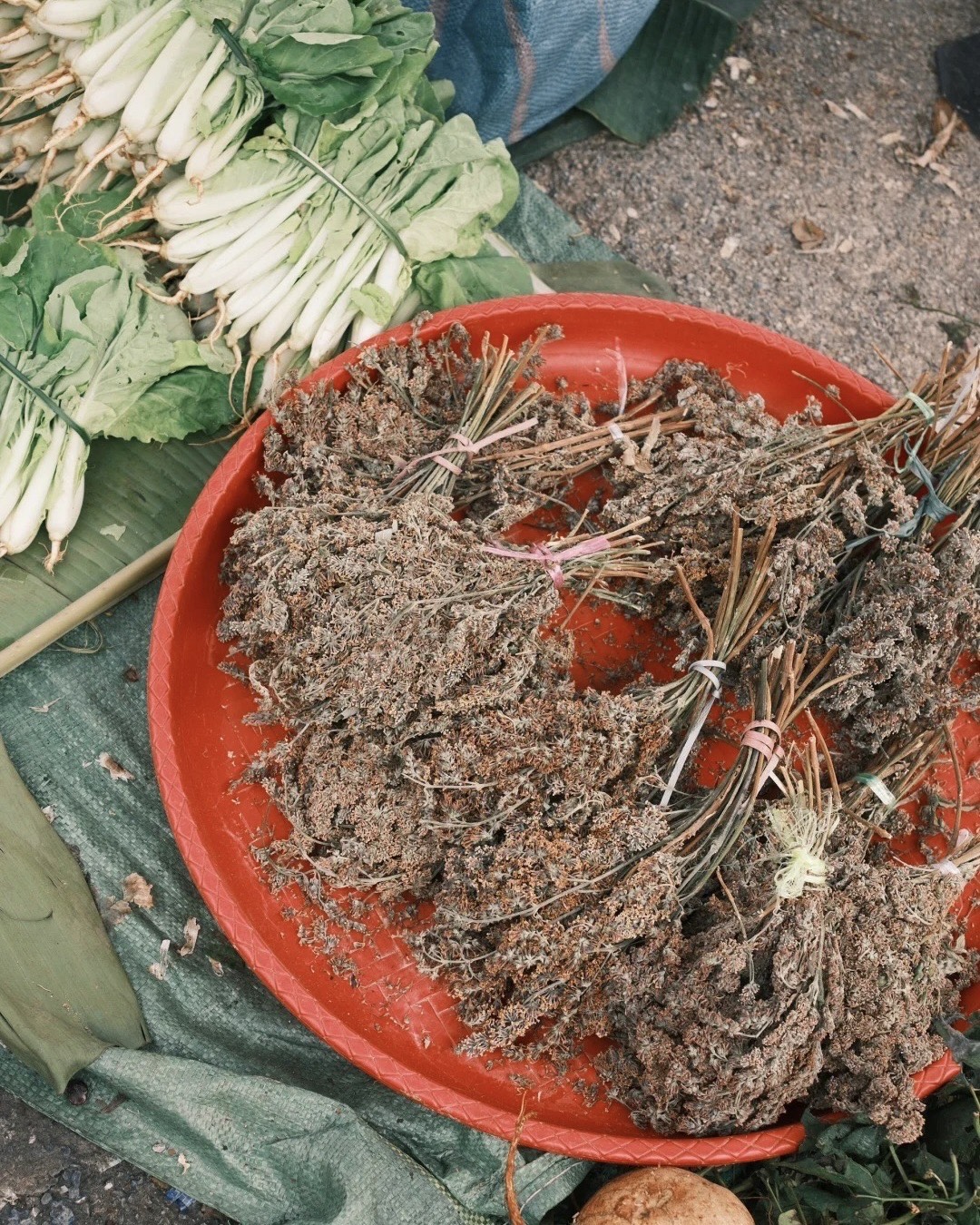 Bundles of dried herbs on a red tray beside fresh bok choy at a Kunming street food market.