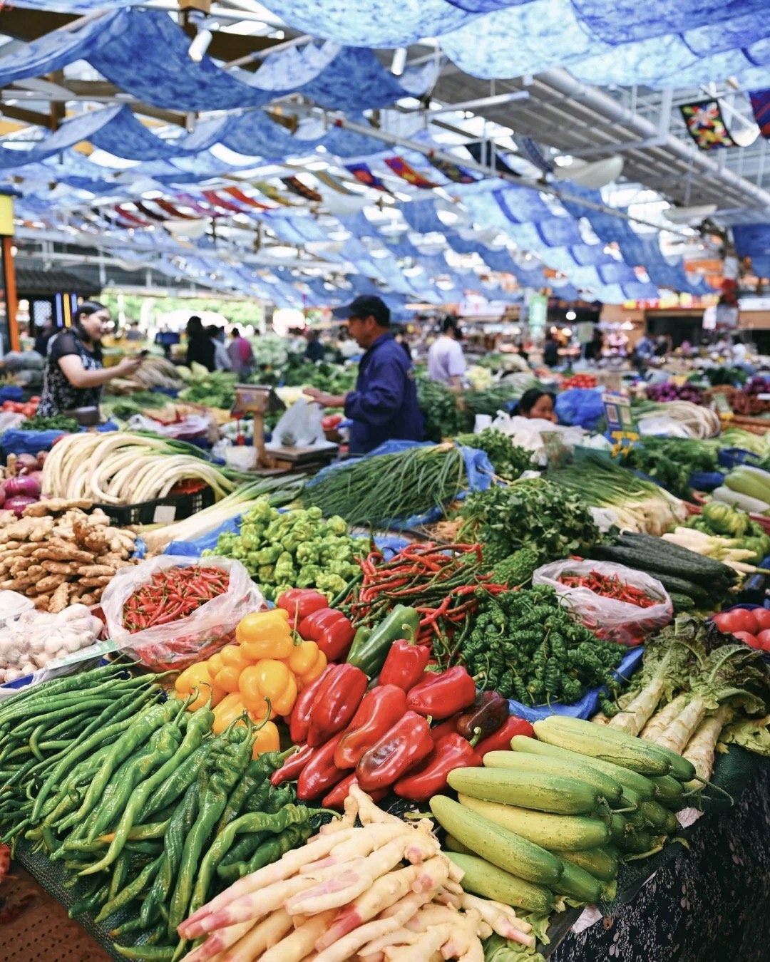 Bustling Kunming food market stall heaped with fresh chillies, peppers, ginger and vegetables under blue canopies.