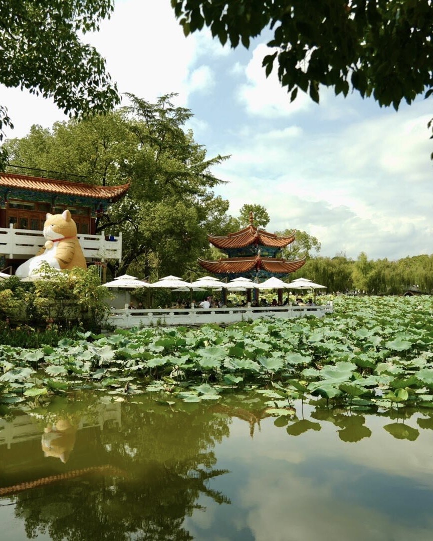 Ornate lakeside pavilions and a giant cat statue overlook a lotus-covered pond in a Kunming city park.