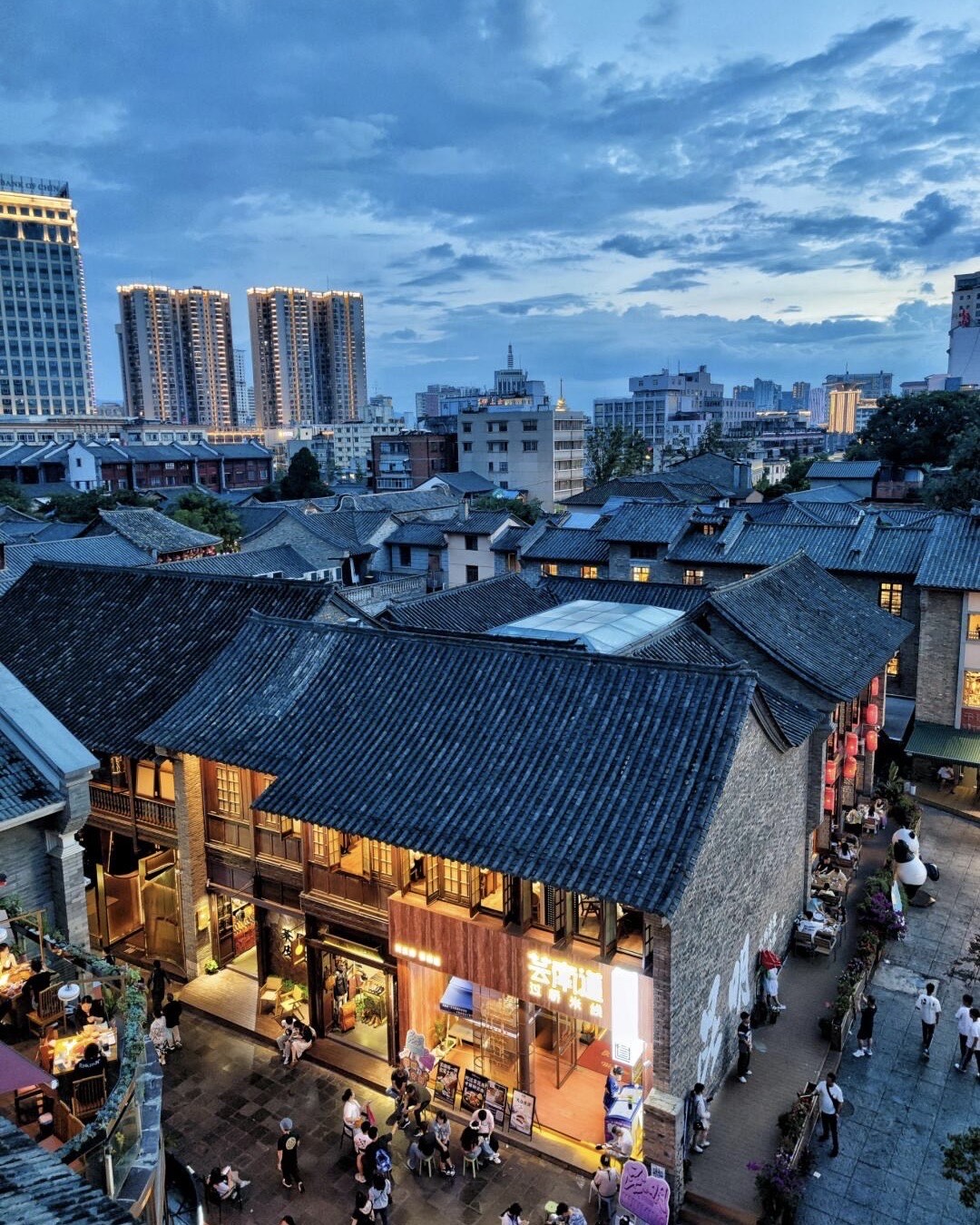 Grey-tiled traditional rooftops of a Kunming heritage quarter glowing at dusk against the modern city skyline.