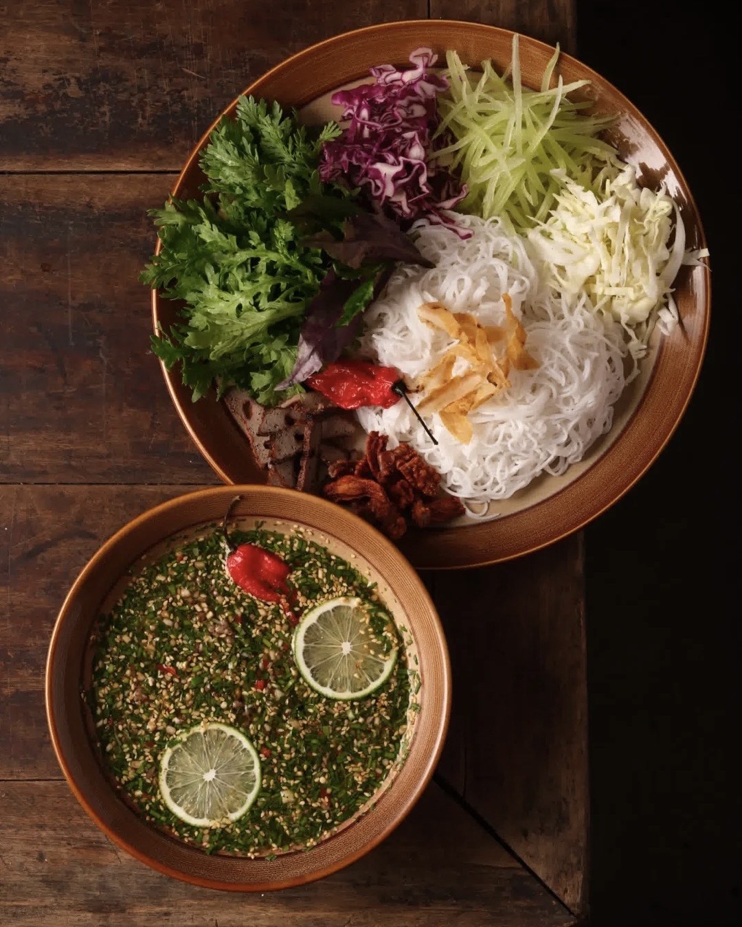 Overhead view of a Kunming rice noodle dish with fresh herbs, shredded vegetables and a lime-topped dipping bowl on a wooden table.