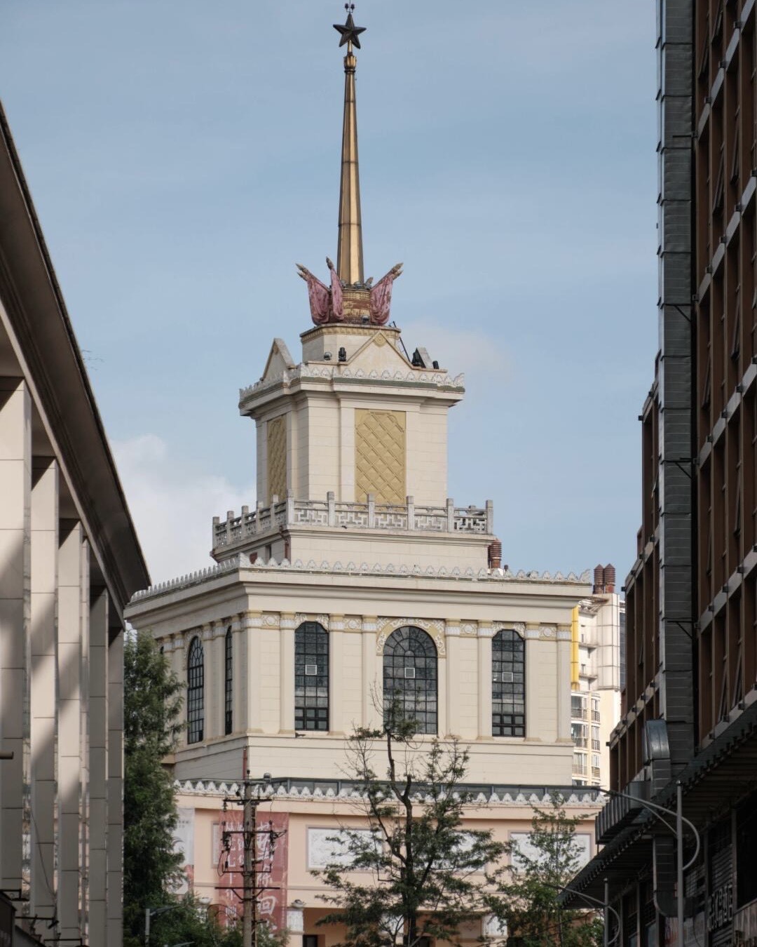 Cream Soviet-era tower with a golden spire and red star rises above Kunming rooftops against a blue sky.