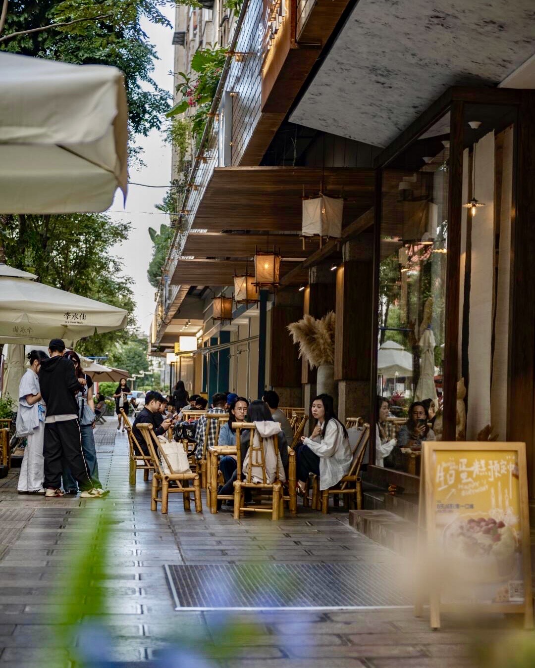 Diners relax in bamboo chairs at a leafy Kunming street cafe with paper lanterns and outdoor umbrellas.