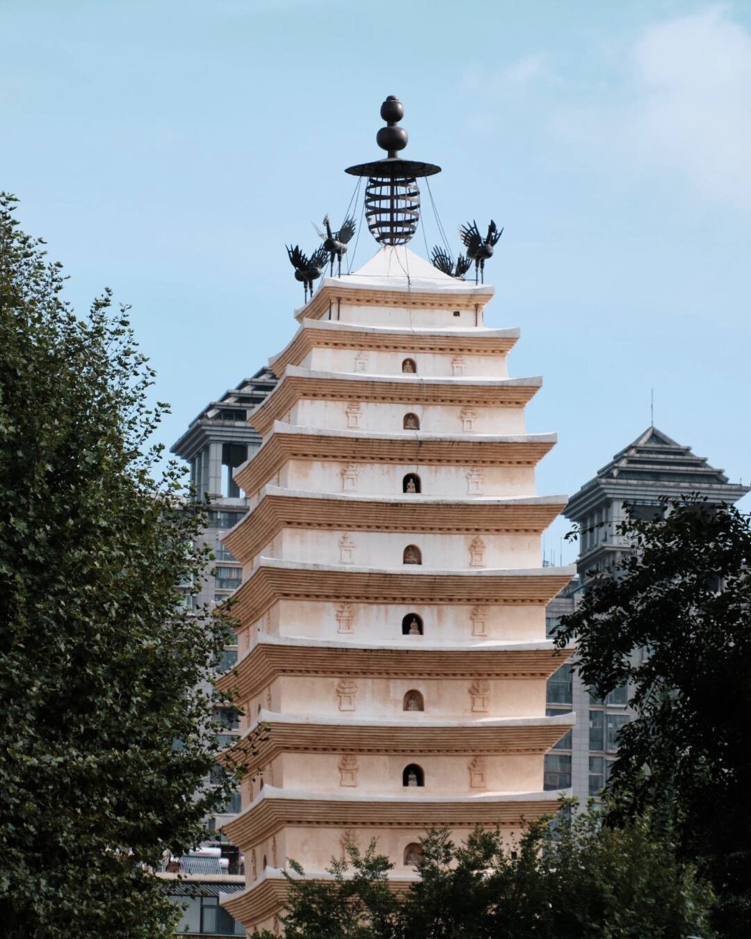 Tall white multi-tiered pagoda topped with bronze bird sculptures rising above trees and modern city buildings in Kunming.