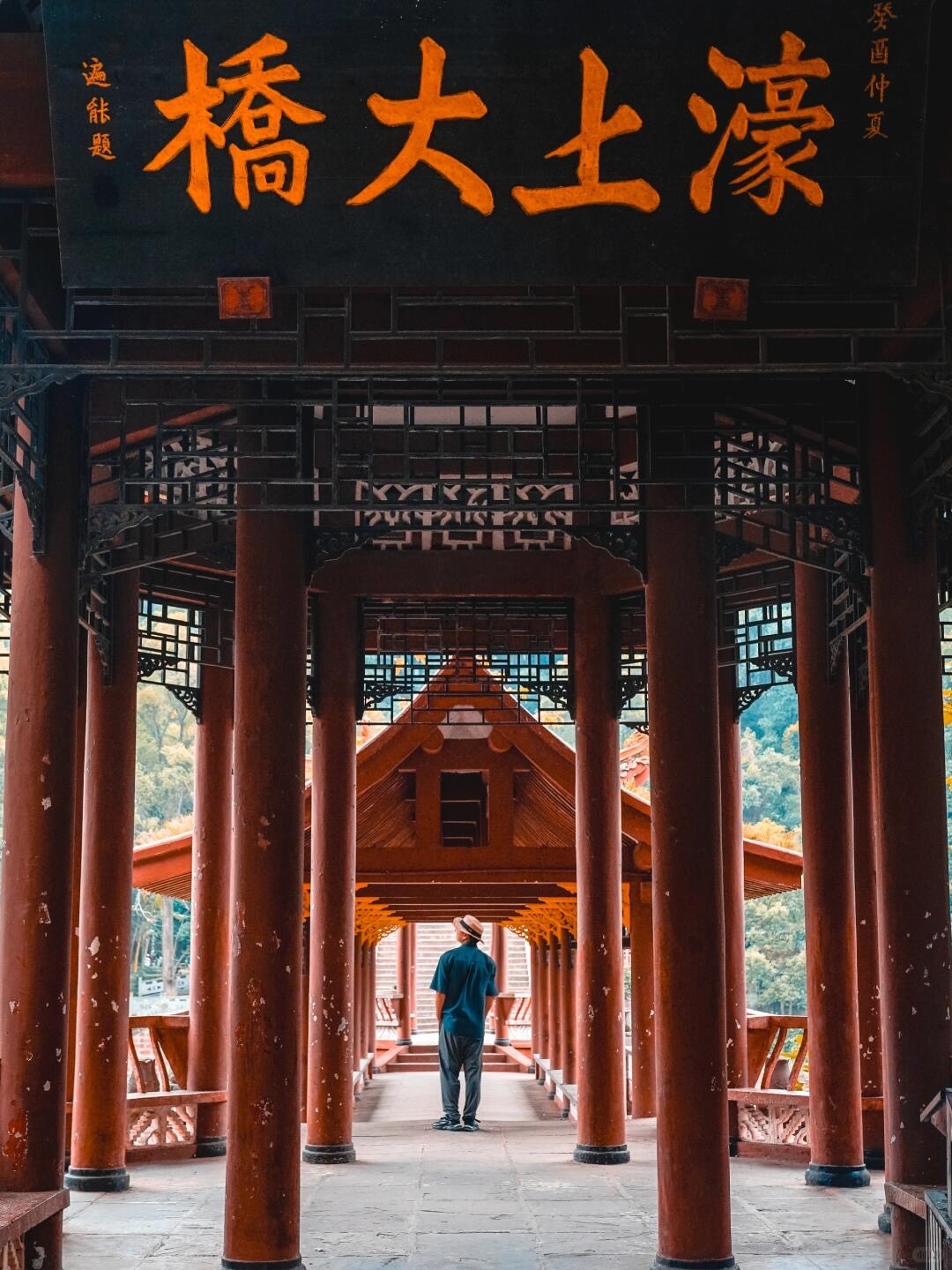 Traveller standing beneath the red timber columns of a covered bridge with a gilded Chinese calligraphy plaque near Leshan.
