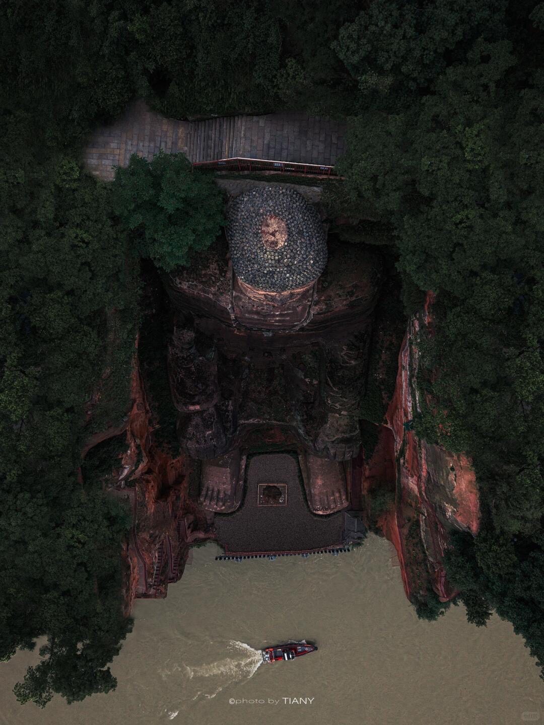 Aerial top-down view of the Leshan Giant Buddha carved into a red cliff as a boat passes on the river below.