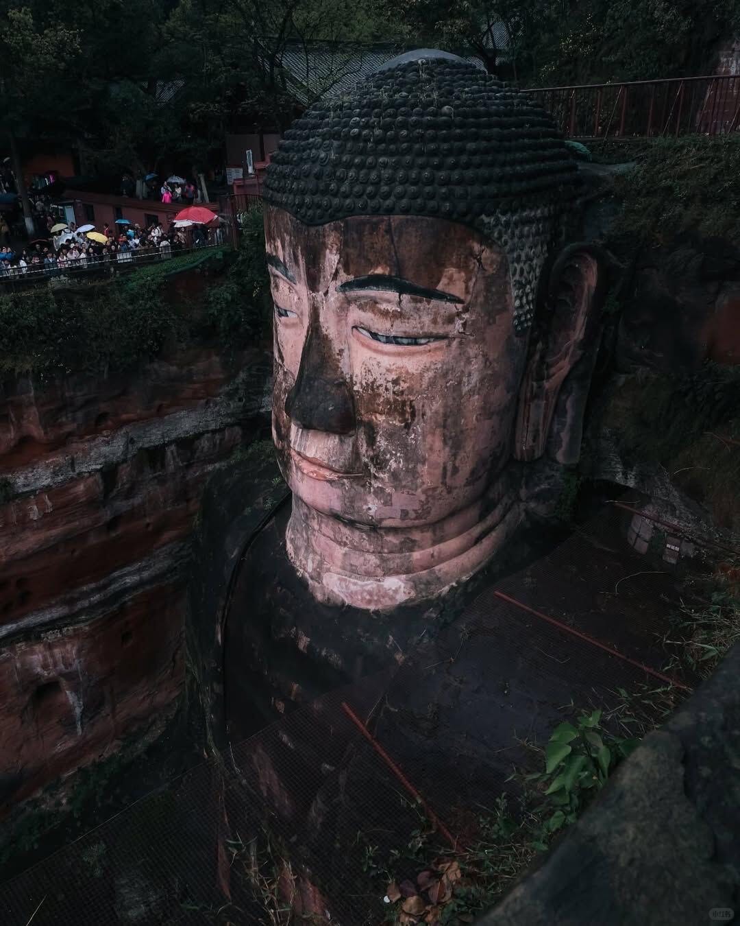 Close-up of the weathered stone face of the Leshan Giant Buddha with visitors on the cliff walkway above.