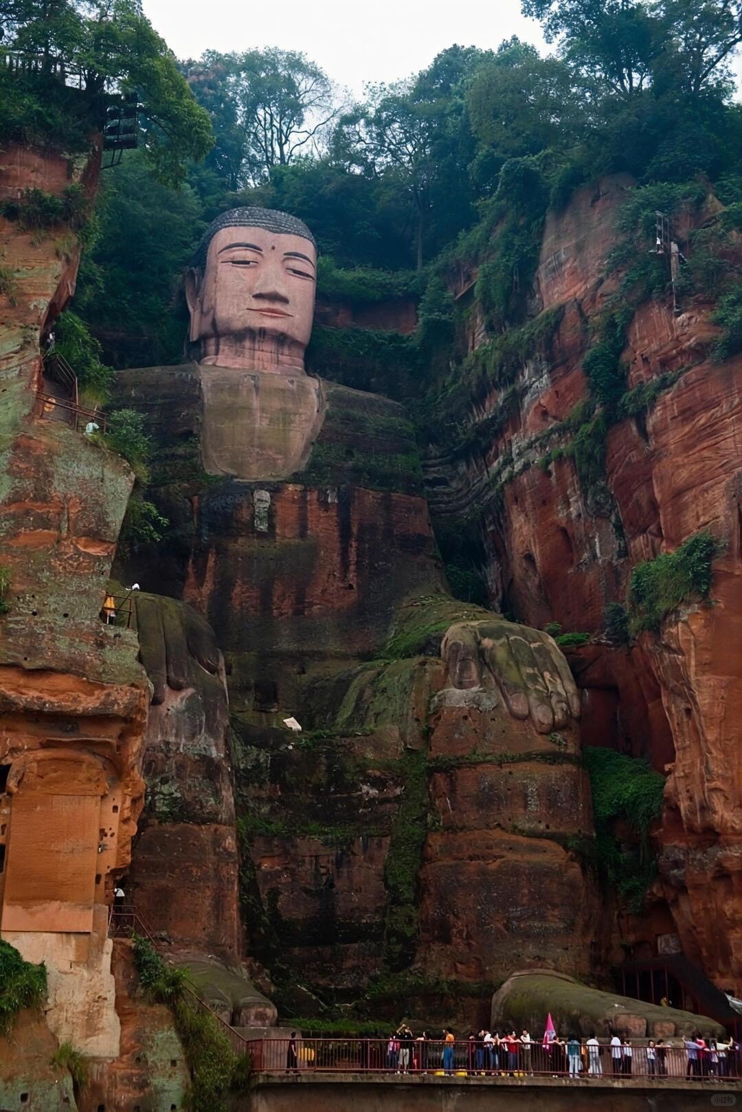 The towering Leshan Giant Buddha carved into a red sandstone cliff with tiny visitors at its feet.