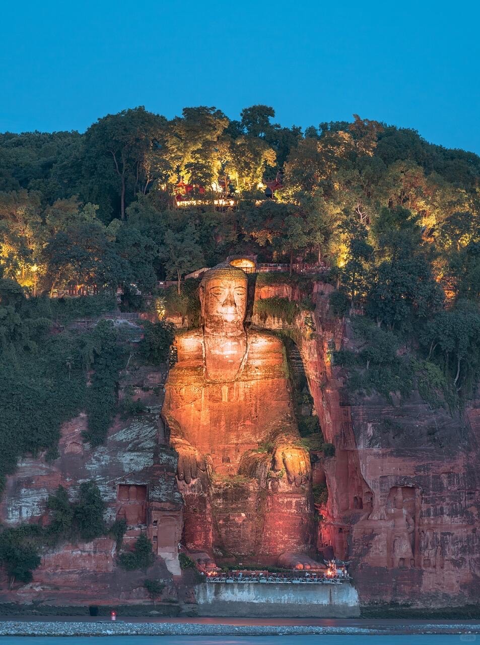 The Leshan Giant Buddha carved into a red cliff above the river, lit against a deep blue dusk sky.