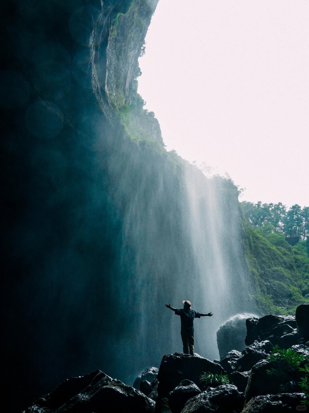 A lone figure with arms outstretched stands on rocks beneath a tall waterfall spilling over a green cliff near Leshan.