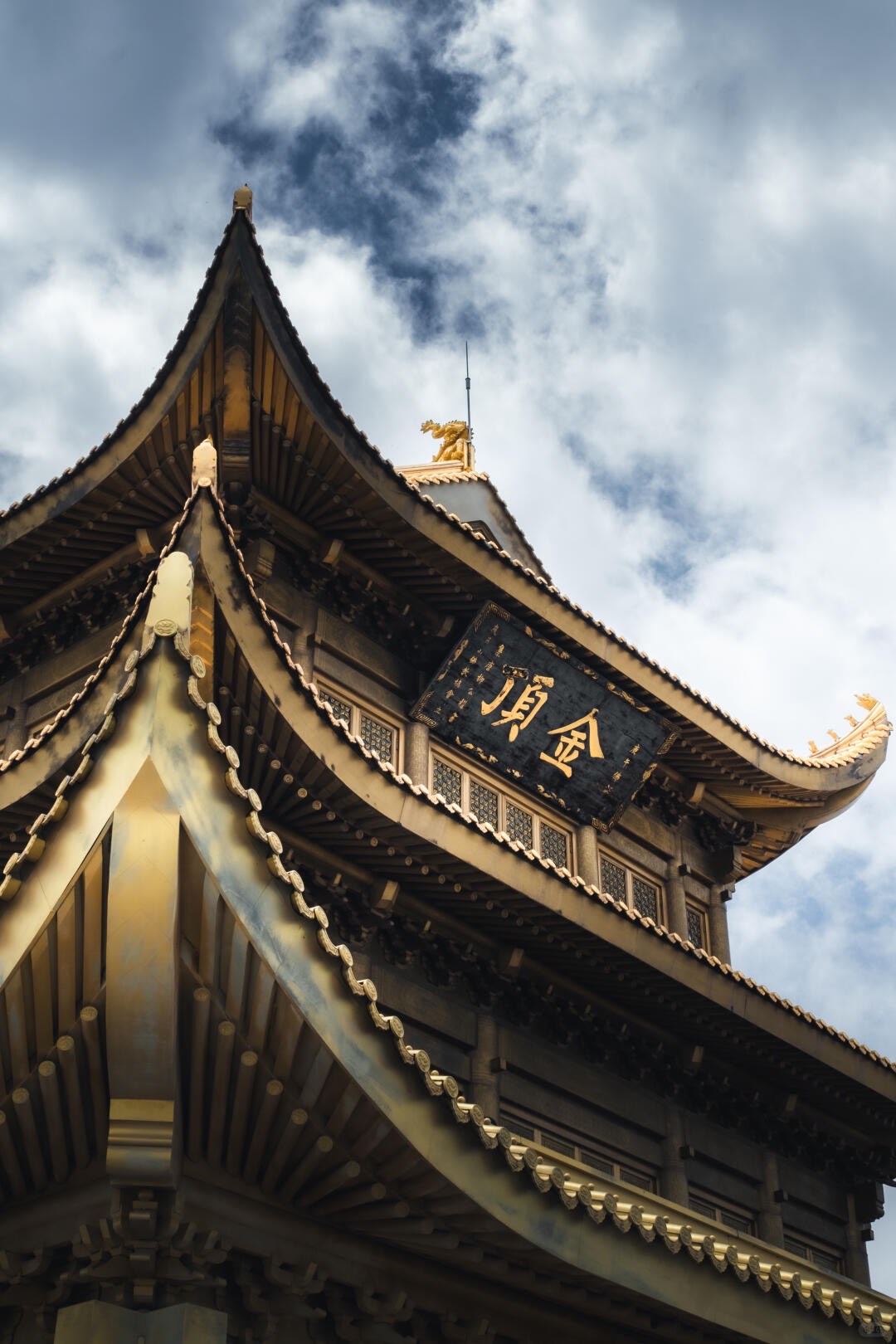 Upswept golden temple eaves and dragon finial of Mount Emei's Golden Summit hall against a cloudy sky.
