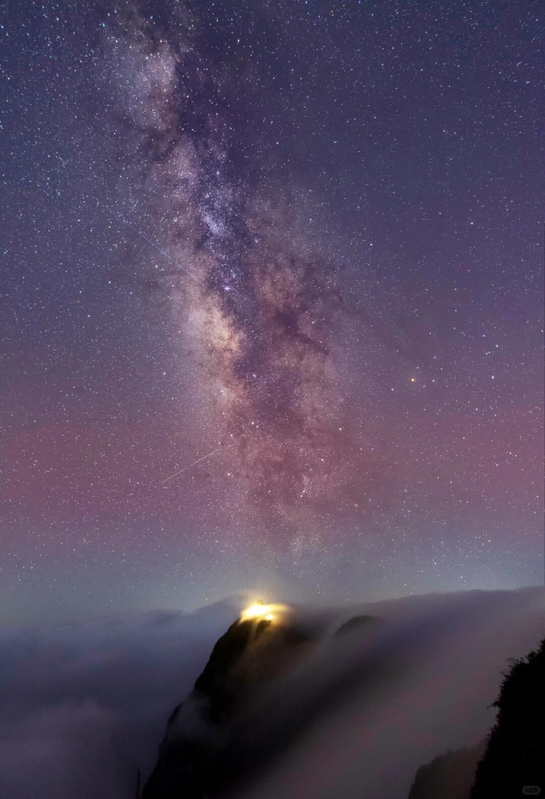 Milky Way arcing over Mount Emei's Golden Summit as a warm light glows through the cloud sea at night.