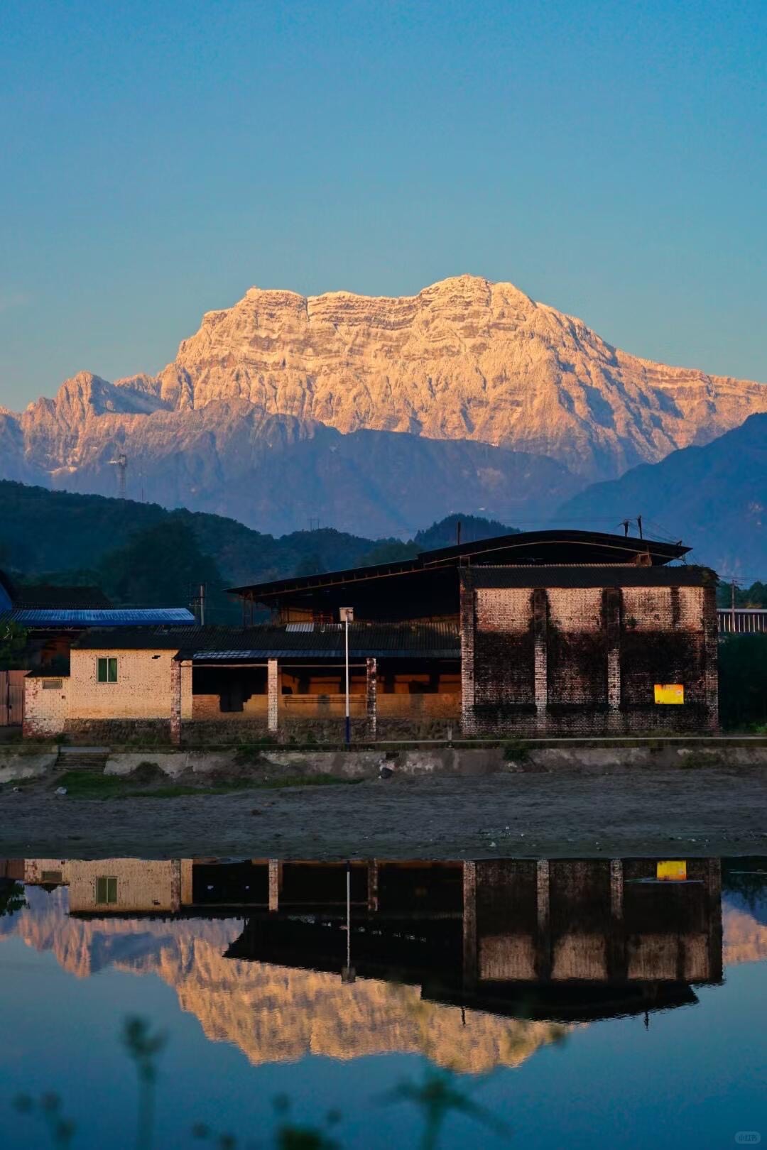 Sunlit rocky peak near Mount Emei reflected in a still pond behind a riverside village at dawn.