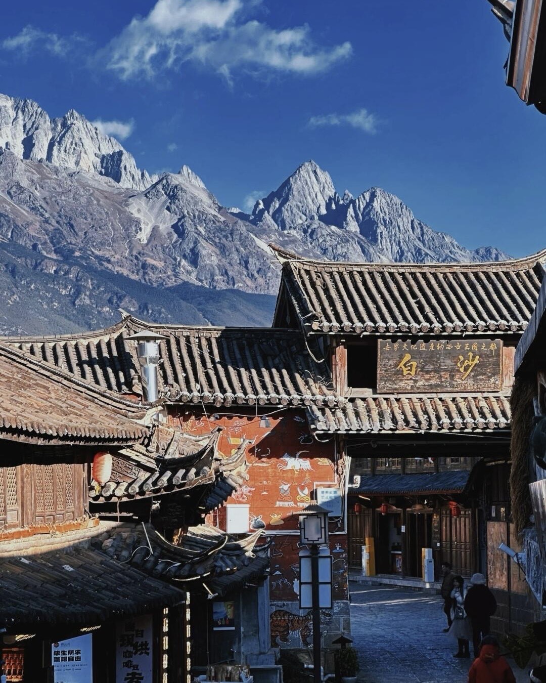 Baisha old town gate and traditional tiled rooftops set against the jagged snow-capped peaks of Jade Dragon Snow Mountain.