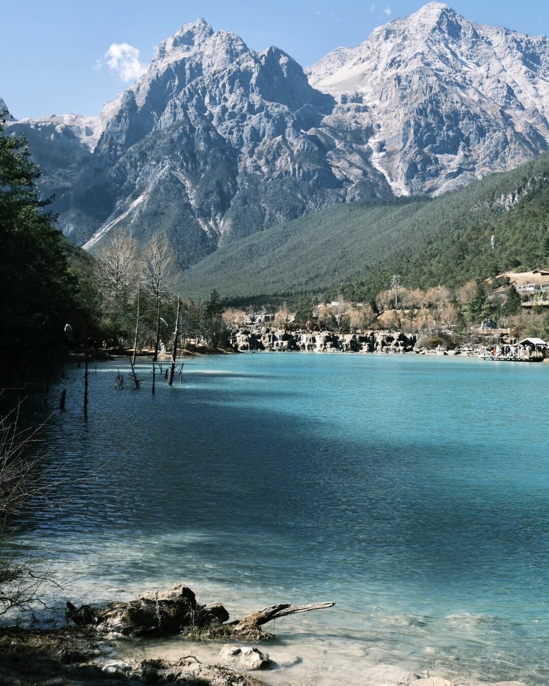 Turquoise blue lake with cascades below the snow-dusted peaks of Jade Dragon Snow Mountain and pine-forested slopes.