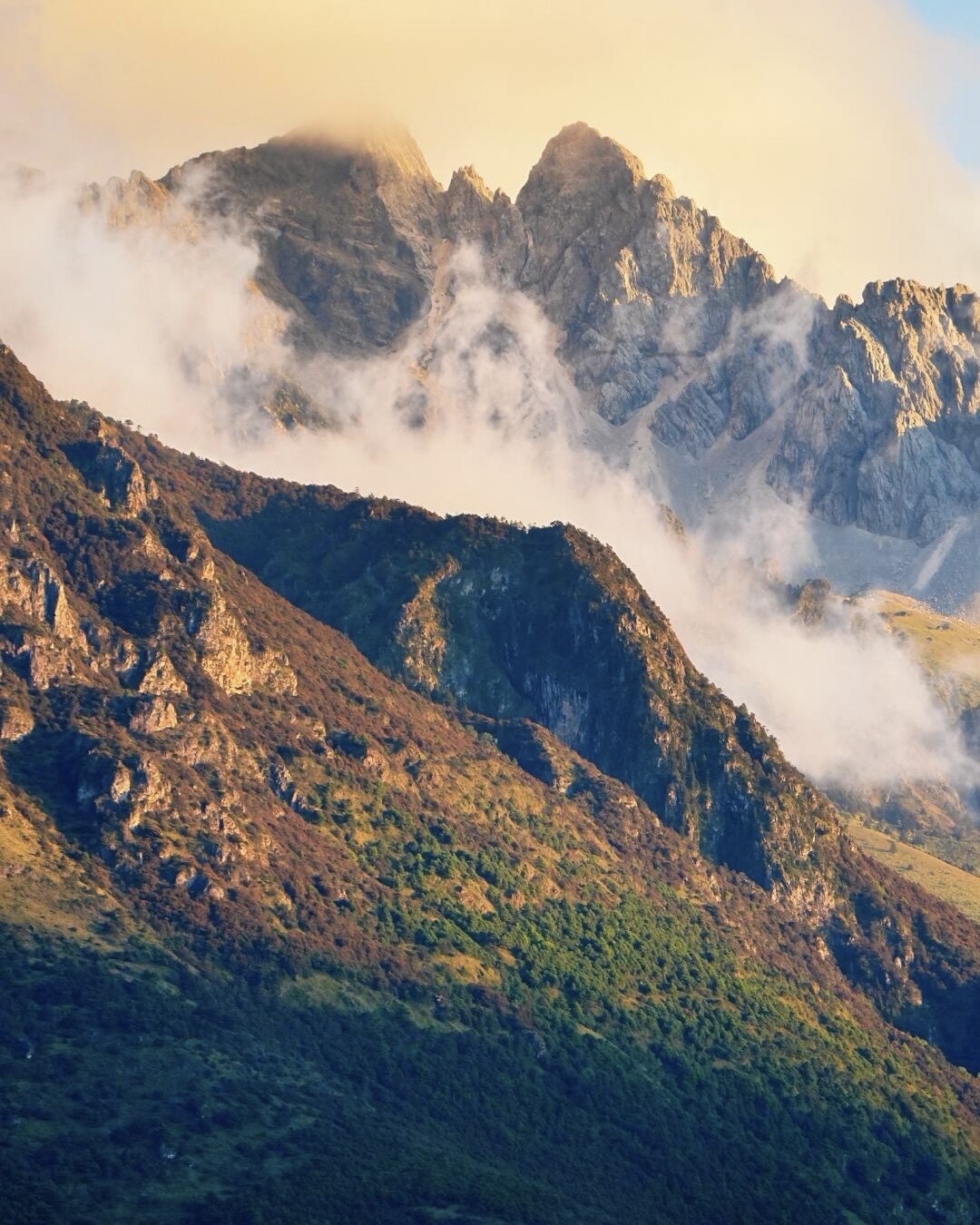 Craggy sunlit ridge of Jade Dragon Snow Mountain wreathed in drifting mist above forested slopes.