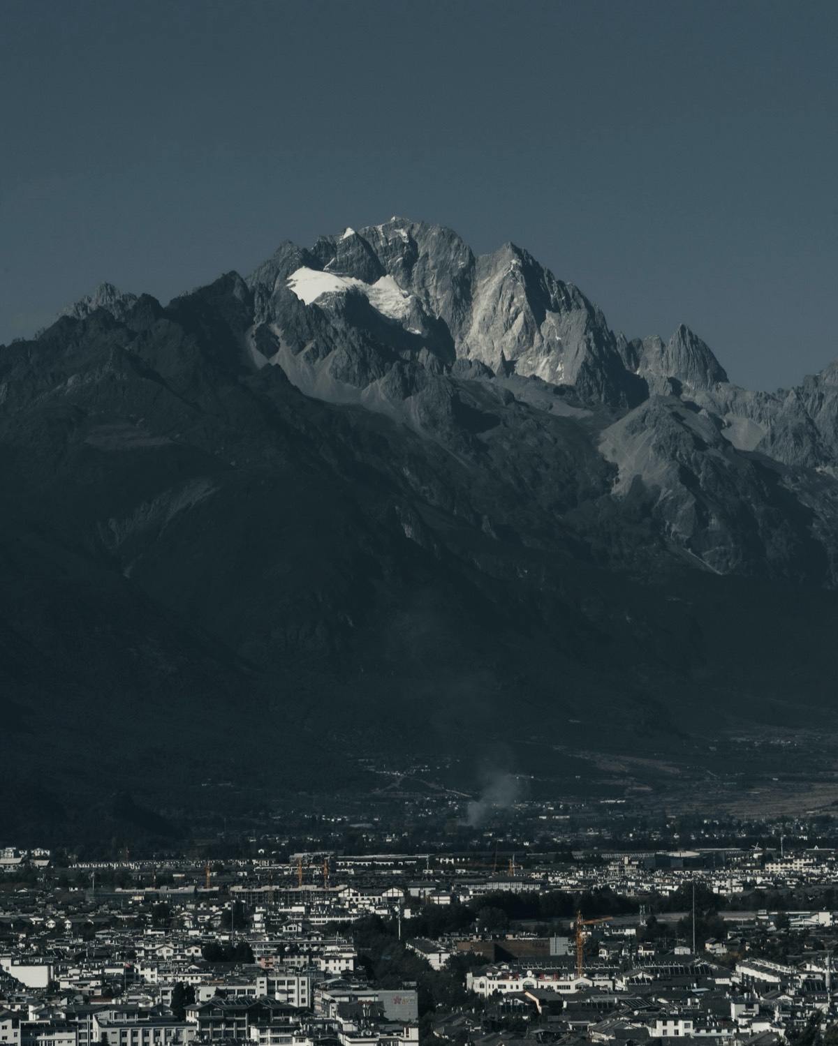 Snow-capped Jade Dragon Snow Mountain rising above the low rooftops of Lijiang city under a clear dark-blue sky.
