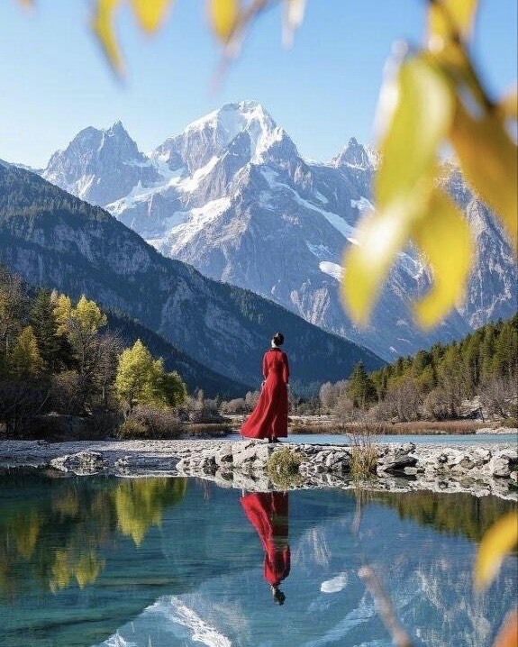 Woman in a red dress by a still lake reflecting snow-capped peaks framed by autumn leaves near Lijiang.