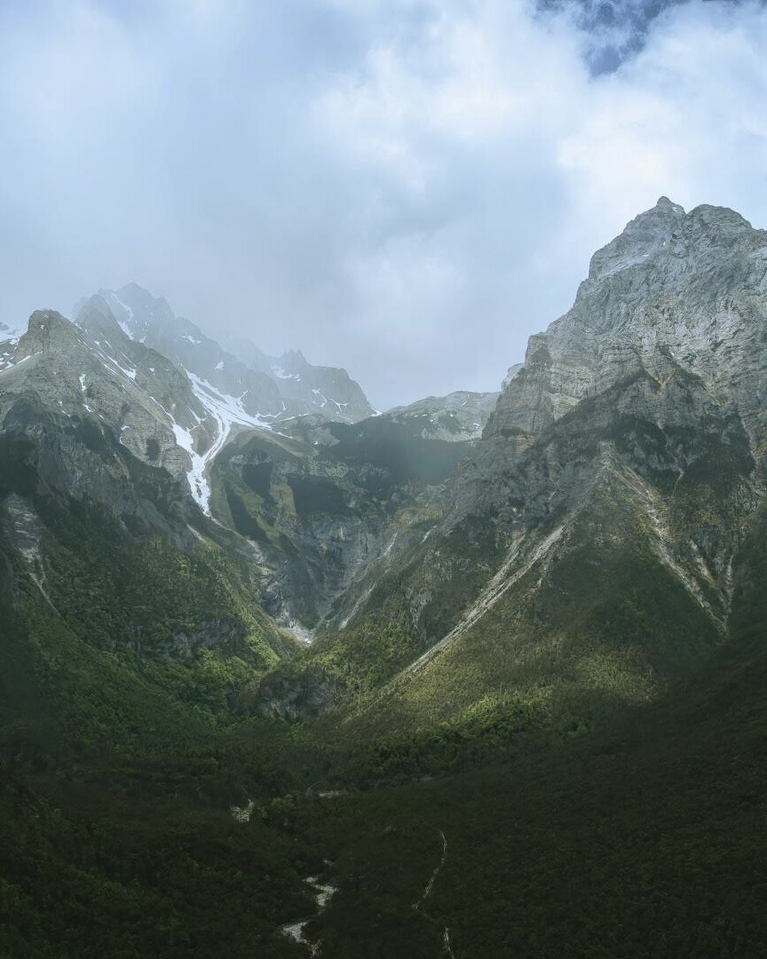 Craggy grey peaks of Jade Dragon Snow Mountain rising above a green forested valley under cloudy skies.