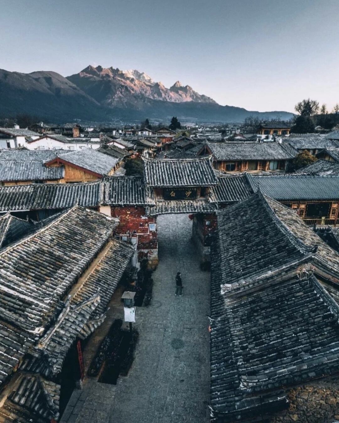 Aerial view over Baisha old town's grey-tiled rooftops and gateway with a snow-capped mountain rising behind at dusk.