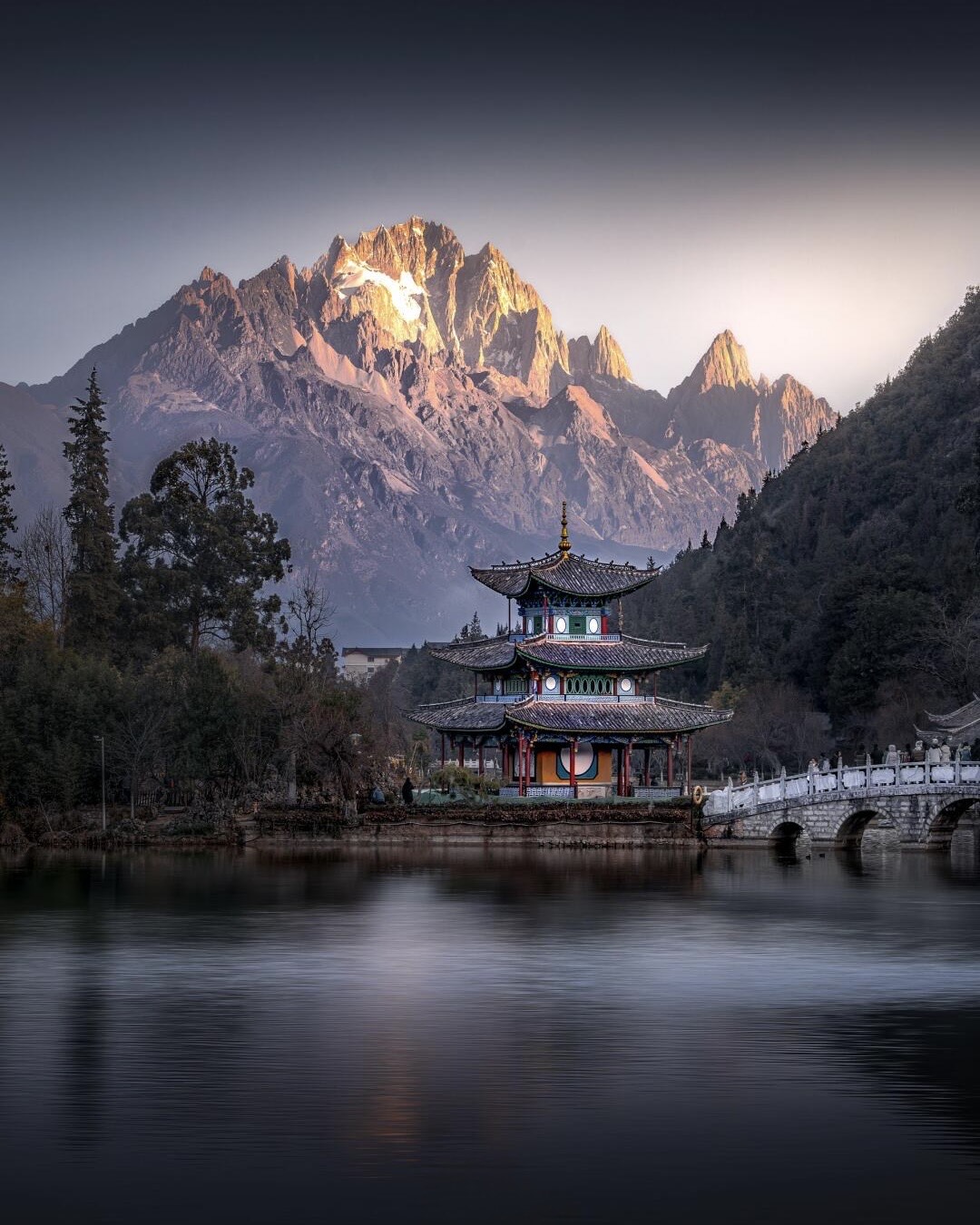 Tiered pavilion and stone arch bridge reflected in a still pool below snow-capped peaks at Lijiang.