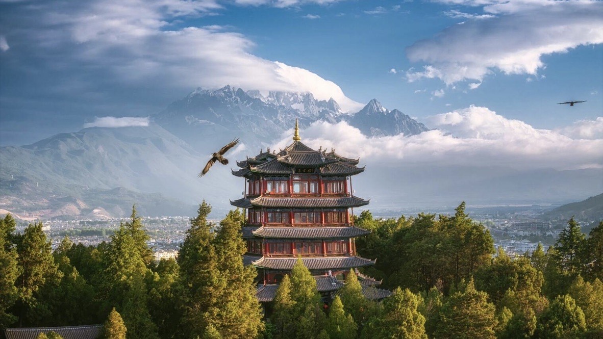 Multi-tiered pagoda rising from pine forest with eagles soaring before snow-capped mountains near Lijiang.