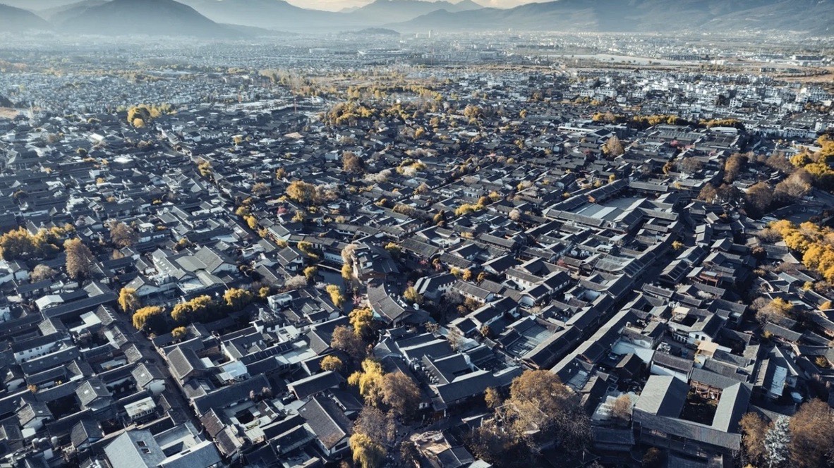 Aerial view over the grey-tiled rooftops of Lijiang Old Town spreading toward distant mountains.
