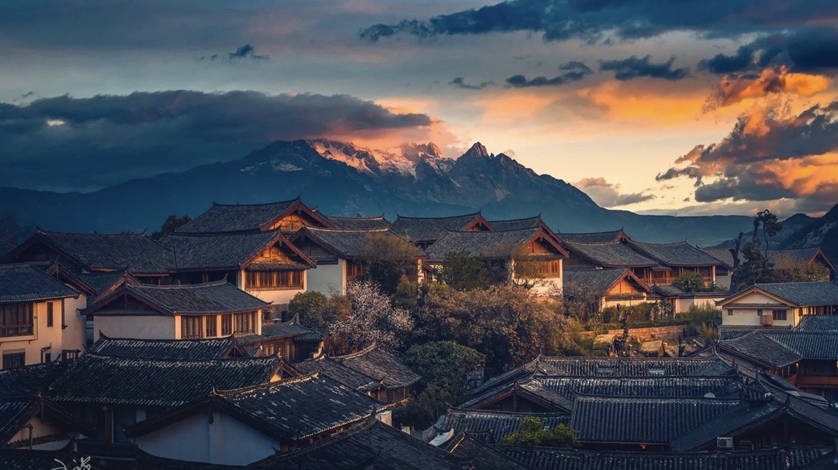 Tiered wooden rooftops of Lijiang Old Town glowing at sunset beneath a snow-capped mountain range and dramatic clouds.