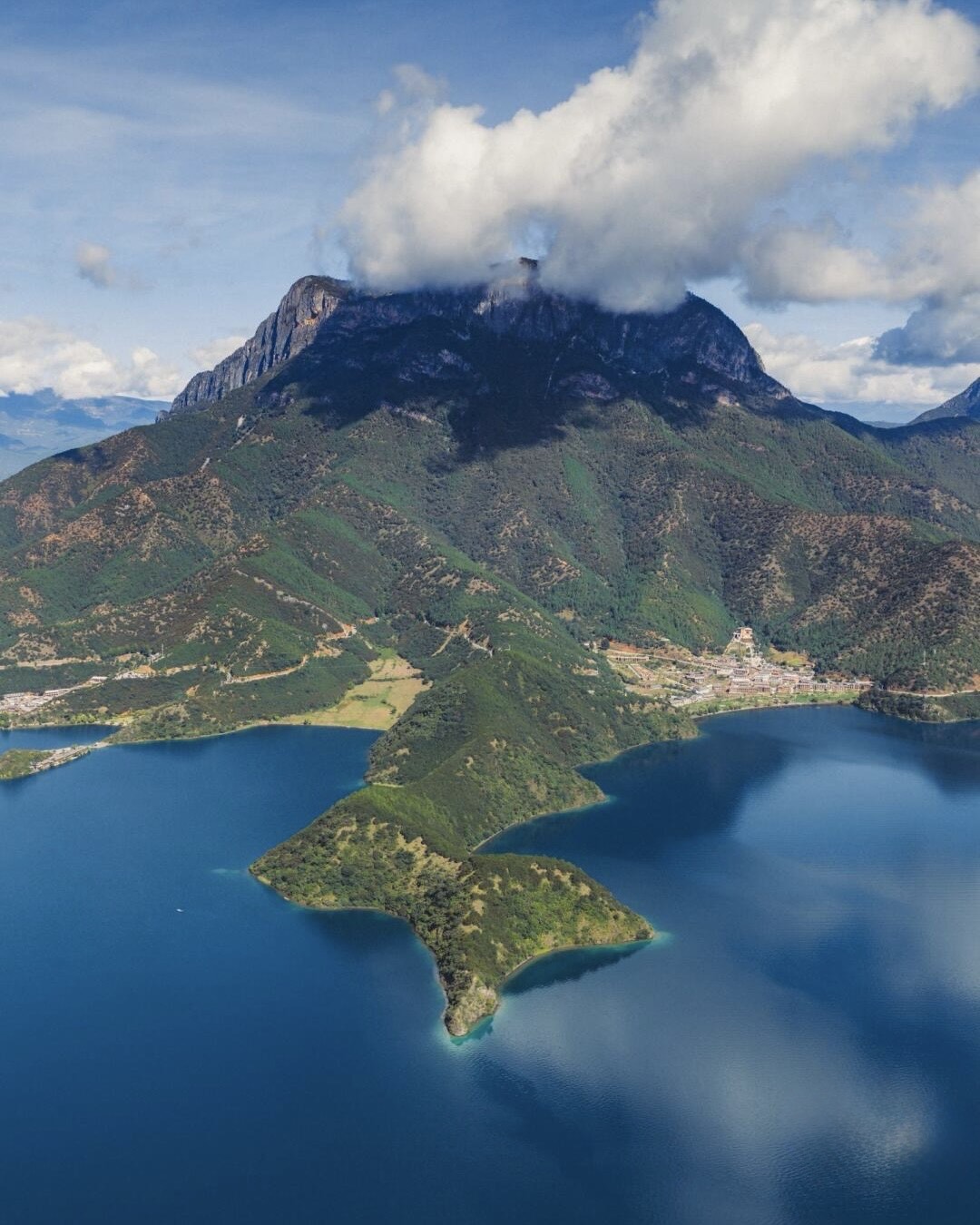 Aerial view of a forested green peninsula reaching into deep-blue Lugu Lake beneath a tall cloud-topped mountain.