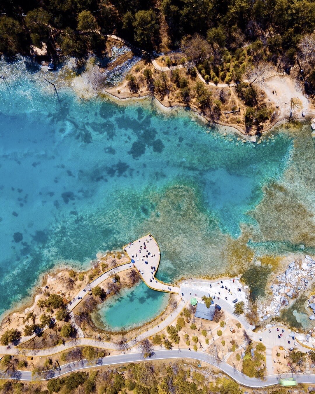 Overhead view of Lugu Lake's turquoise shallows with a wooden boardwalk and pier lined by visitors on the shore.