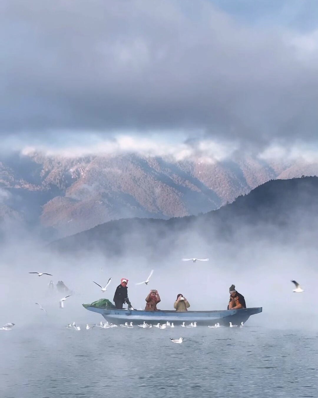 People in a wooden boat feeding gulls on misty Lugu Lake with snow-dusted mountains rising behind.