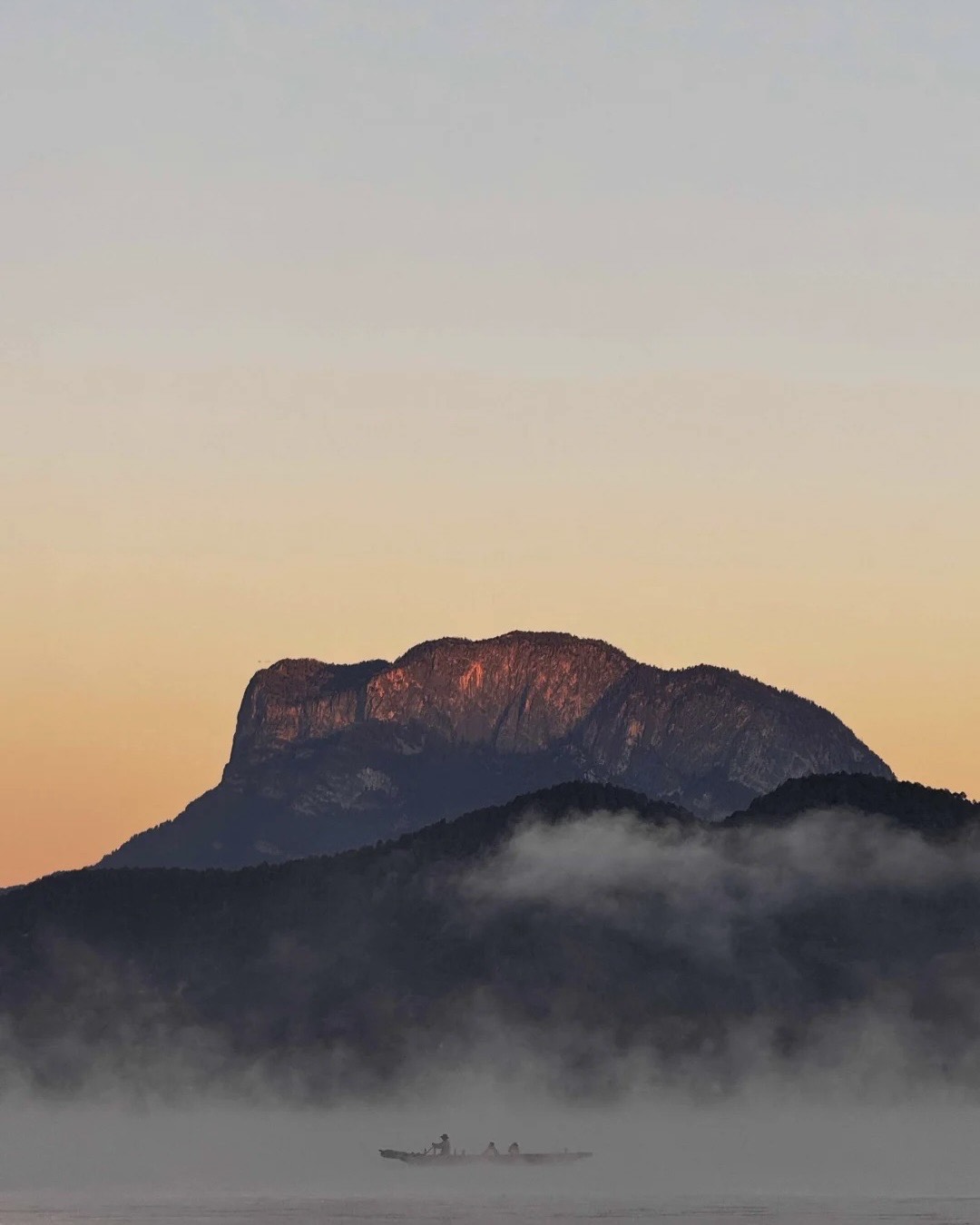 Silhouetted boat drifting through low mist at sunrise beneath a mountain glowing in warm dawn light near Lugu Lake.